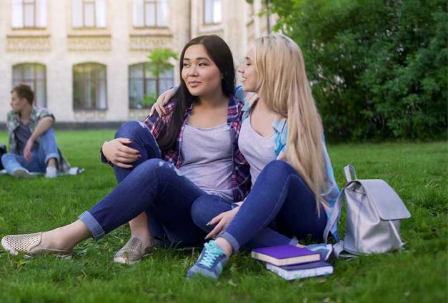 Two female friends sitting on lawn near college, hugging