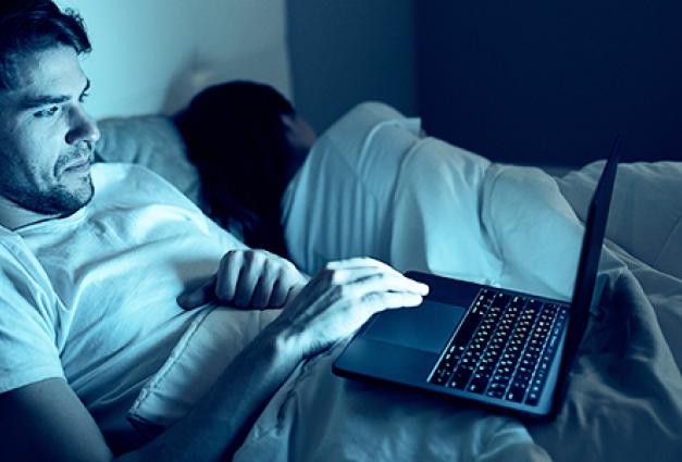 Young man on his laptop in bed next to his partner who is sleeping.