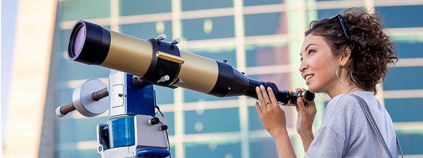 A woman looks across a telescope in front of a glass building