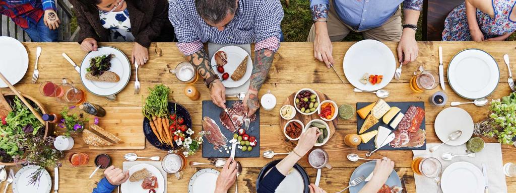 group of people eating otuside at a big table, some are men, some are women, young and old. boards of food and shared plates show people eating.
