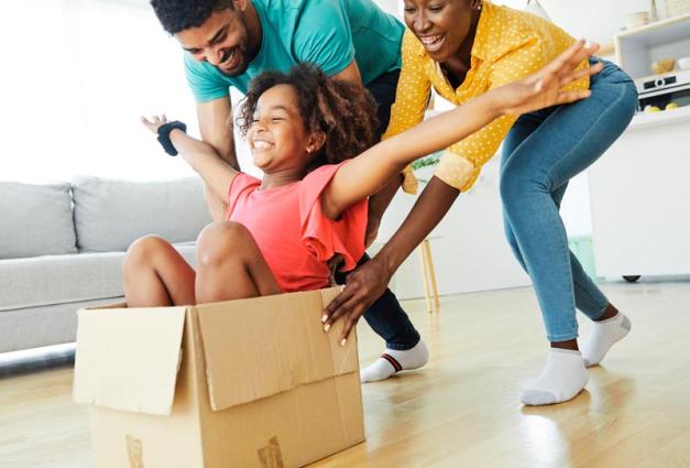 Father and mother pushing young daughter along floor in cardboard box