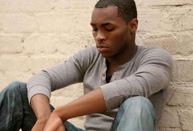Young Black man sitting leaning against a brick wall looking down