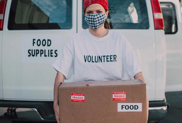 Young woman wearing a mask and volunteer shirt holding a large box of food supplies