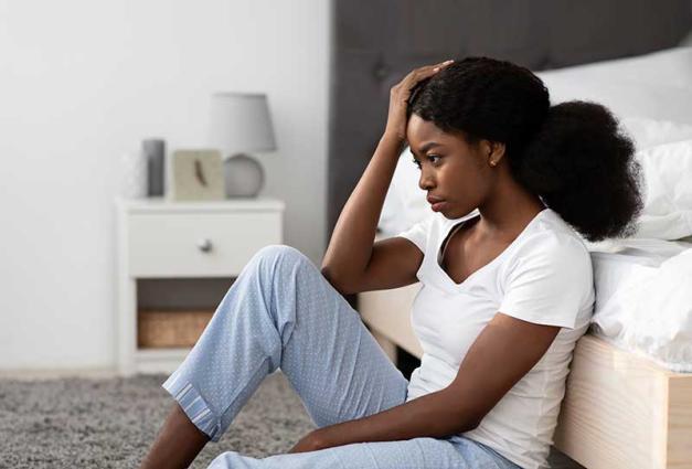 Stressed young black woman sitting on floor by bed at home