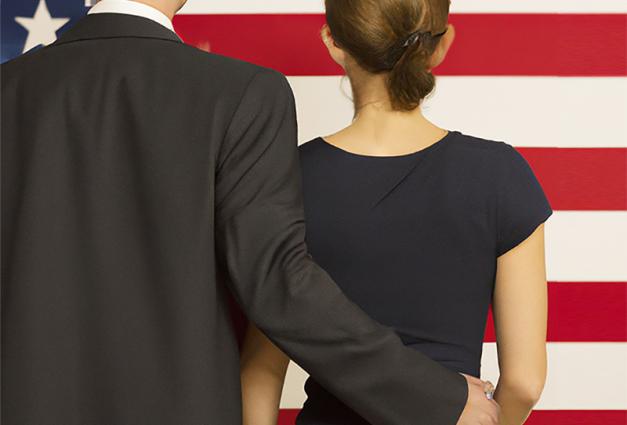 man and woman standing in front of a flag. Man's hand is on woman's lower back.