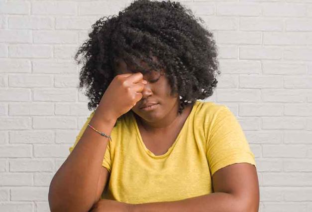 Black woman sitting in front of a white brick wall rubbing her eyes