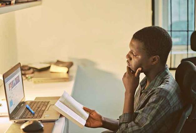 man reading book while sitting in front of computer