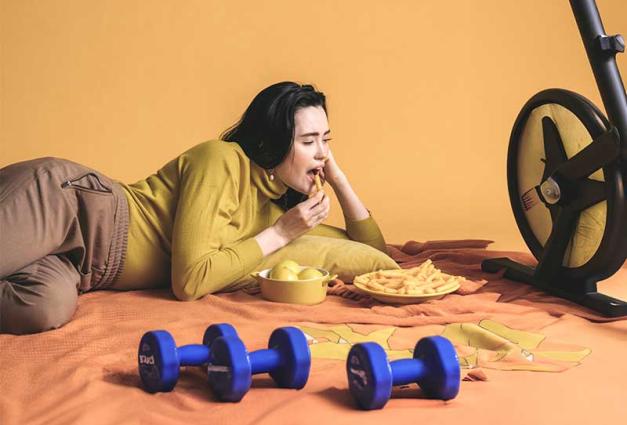 Woman lying on floor next to dumbbells and exercise bike eating french fries