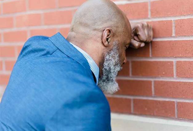 Older Man wearing a blus suit leaning his head against a brick wall