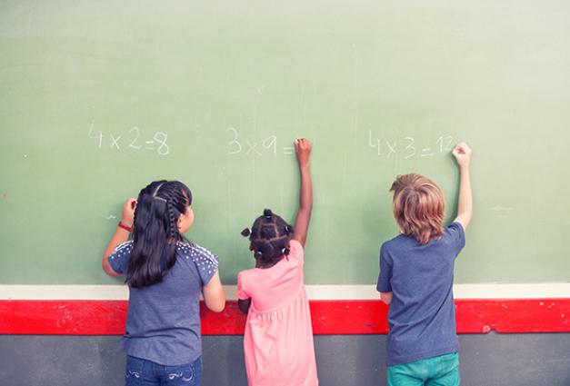 Three young children solving multiplication problems on a chalkboard