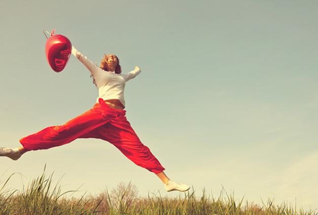 Woman with red heart balloon in her hand jumping in the air over a green field