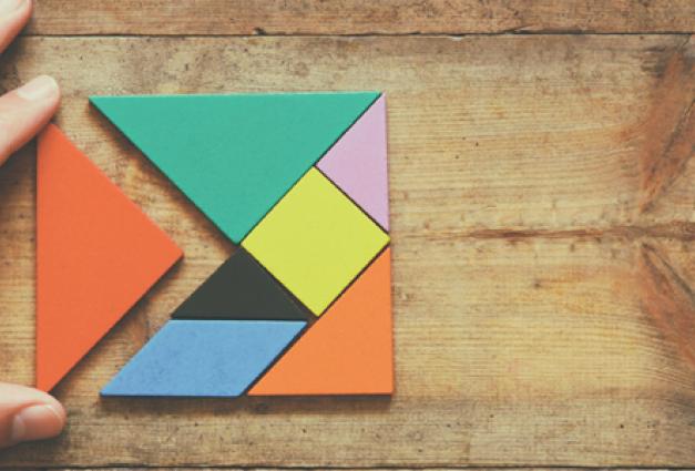 man's hand holding a missing piece in a square tangram puzzle, over wooden table.