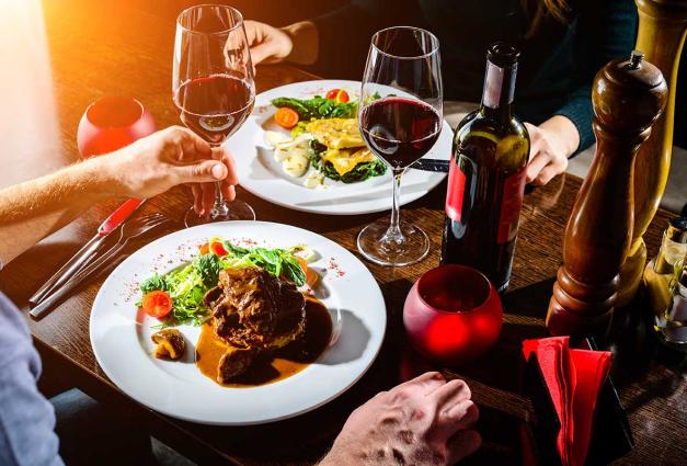 Image of a table with w full plates of food, two wine glasses with red wine, and a couple's hands in position to eat