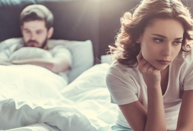 Image of woman sitting on the edge of the bed, upset/thinking, with man frustrated laying in bed